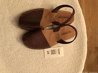 Brown Solillas leather sandals with slingback heel strap, square toe, white stitching on the sole, lying on a light-colored fabric surface.