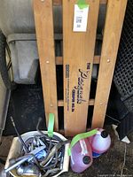 Wooden creeper bench standing vertically with visible 'The Passgate Creeper No 36' branding, two plastic containers of RV antifreeze beside it, and a white tub filled with assorted metal tools and hoses.