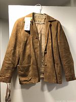 Front view of 1960s vintage ladies suede jacket hanging on a white wall with a size tag 27 visible on the hanger.
