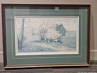 Framed print showing three cows sitting under blossomed trees in a pastoral setting with a fence and distant background, featuring a green mat and dark wood frame.
