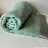 Rolled celadon green wool blanket showing fabric texture and satin trim.