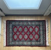 Rectangular wool rug in red with turquoise border and fringe along the short edges, placed on a hardwood floor near a door, showing the full rug view.