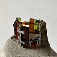 Close-up view of the bracelet showing the colorful resin panels in amber, olive green, yellow, and brown hues, accented by textured pewter geometric shapes on an elastic band.