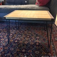 Full front view of the charcuterie tray table placed on a patterned rug in front of a gray couch, showing wooden tray top and black thin hairpin legs.