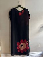 Front view of the black sheer dress with bold red and pink poppy flower print near the bottom front hem, hanging on a hanger against a wall.