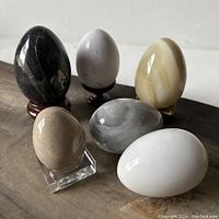 Six polished eggs arranged on a wooden surface, showing various colors and textures from black and gray to beige and white, with a selection of four different stands visible beneath some eggs.
