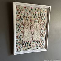 Framed embroidered artwork showing peace and love hand signs with floral background from an angled side view.