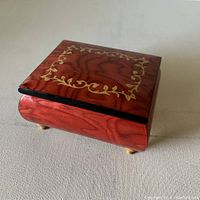 Wooden rectangular Sorrento music box showing detailed gold floral inlay and reddish wood grain finish, resting on small feet.