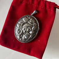 Front view of the oval sterling silver locket showing floral repousse design resting on red velvet pouch.