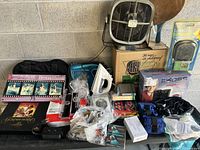 Photo showing a houseware lot with a white steam iron, box fan, Boss amplifier, assorted costume jewelry, puzzle, and various items in plastic bags.