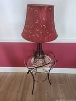 Photo showing the red fabric lampshade with gold swirl patterns on a metal Eiffel Tower base, placed on a circular glass-topped metal bedside table against a red and white wall.