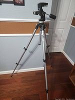 Silver metal Bogen Professional tripod set up on a hardwood floor against a gray and brown wall background providing a view of the entire tripod with its adjustable legs and black mounting head with handle.
