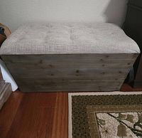 Front view of rectangular wooden chest with beige fabric cushioned top showing natural wood grain and tufted cushion detail.