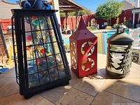 Photo of three lanterns outdoors on tiled surface: large black metal and glass lantern with grid, red metal Christmas lantern with star and moon cutouts, green metal Christmas lantern with embossed tree designs.