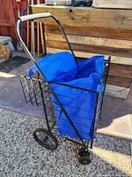 Folding metal wireframe shopping cart with two wheels and royal blue fabric bag inside, shown in outdoor patio area.