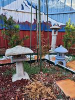 Photo of three Japanese stone pagoda garden decorations outdoors, situated on mulch with garden plants and a painted fence in the background.