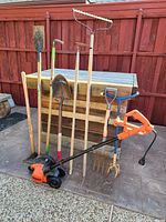 Photo of several garden hand tools propped against wooden crate, including shovels, fork, rakes, edger.