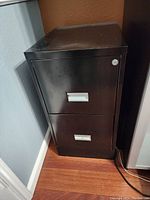 Black metal filing cabinet with two drawers placed against a wall on wooden floor.