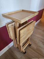 Four individual wooden folding party tables stacked together with tray holders visible. The tables have light wood finish, raised edges to hold items, foldable legs, and wheels on bottom for movement.