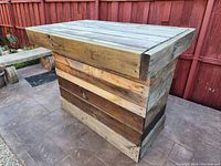 Photo of a wooden patio bar table outdoors on a paved surface with a wooden fence in the background. The table is made from different wood panels in natural and darker tones.