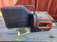 Powershot Pro electric staple gun, black and orange, placed on wooden surface next to its black hard plastic carrying case and a pack of staples.