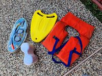 Five snorkeling related items including red life jacket, snorkeling mask with snorkel, yellow swim fin, white peanut-shaped foam float, and blue sponge float on stone ground.