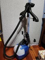 Clothes steamer base, hose, and attached hanger stand shown against a grey fabric backdrop on a hardwood floor.