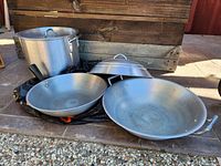 Photo shows one 20 quart stainless steel stockpot with lid and two metal woks of different sizes, one with lid and one without, placed on the ground.