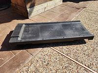 Black hard plastic creeper laid on the ground outdoors, showing the top surface with an elevated headrest area and minor wear marks.