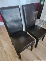 Two black vinyl upholstered chairs with high backs and wooden legs, one chair shows a small crack on the seat upholstery.