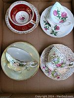 Four assorted porcelain cups and matching saucers arranged in a box, showing varied floral designs and gold trim.