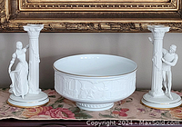 Full view of two white porcelain candlesticks with classical figurines and columns surrounding a white decorative bowl with embossed designs.
