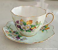 Shows the cup and saucer set from the front, displaying the colorful floral pattern and gold trim accents along scalloped edges.
