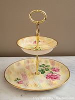 Two-tier ceramic dessert stand with pink rose floral pattern and gold trim, shown from front angle.