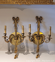 Pair of brass wall sconces mounted on a wall, showing full front view with ornate bows and tassel details.