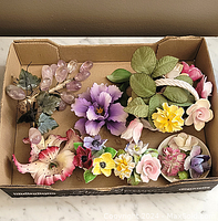 Top view of a cardboard box containing porcelain flower figurines and glass grapes with green leaves.