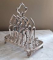 Front angled view of the silver plated toast rack showing ornate handle, curved dividers, and decorative base with floral filigree design.