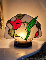 Photo of Tiffany style table lamp lit, showing stained glass shade with red flowers and green leaves on white background on wooden base.