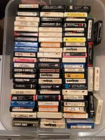 Top-down photo of a plastic bin filled with 54 assorted 8-track stereo tape cartridges with visible artist labels and album titles.