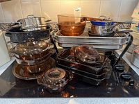 Top view showing a stack of amber CorningWare glass pots with lids, rectangular baking dishes, stainless steel and blue enamel pots, and metal roasting pans on a black stovetop.
