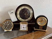 The full lot shown on a wooden surface: one wooden mantel clock and three smaller alarm clocks with different designs and sizes.
