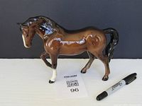 Brown glossy ceramic horse figurine with one white front leg, posed mid-step, shown from front and side angles with Sharpie marker for scale and maxsold lot tag visible.