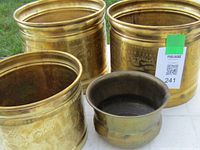 Three large cylindrical brass planters and one small squat brass pot displayed on white surface outdoors