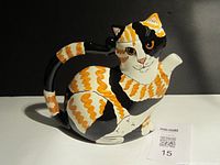 Front view of the calico cat shaped ceramic teapot with a MaxSold card beside it.