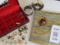 Overview of costume jewelry lot showing rings, silver bracelet, floral brooch, red jewelry box, and beige roll-up organizer