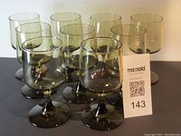 Front view of nine smoke-colored MCM style wine glasses arranged closely together with a MaxSold tag in front showing lot number.