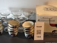 Overall view of 6 clear glass sake glasses and 6 porcelain sake cups with gold rims and portrait designs on a table with a box in background.