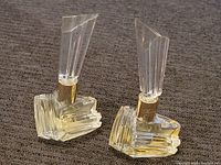Two clear glass Mariah Carey Forever perfume bottles with gold-colored collars and faceted clear caps, each half-full with pale yellow liquid, placed side by side on a dark fabric surface.