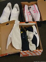 Three pairs of sneakers: Adidas white slip-ons with tags, Under Armour white/pink in box, Nike black/blue Renew In-Season TR 10 in box with paper stuffing.