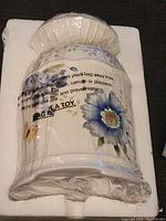 Photo of a white ceramic cookie jar wrapped in plastic, decorated with blue flowers, showing the warning label on the plastic wrap.